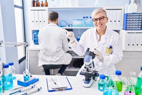 Middle age woman working at scientist laboratory smiling happy pointing with hand and finger to the side