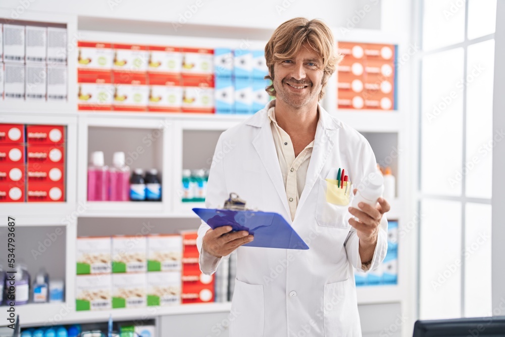 Young man pharmacist holding clipboard and pills bottle at pharmacy