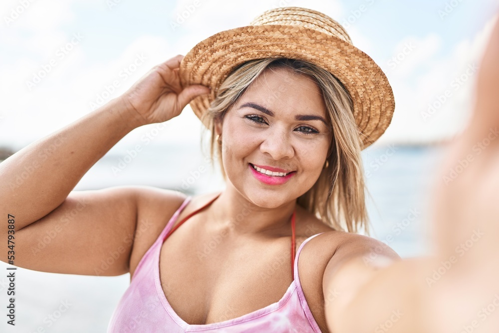 Young hispanic woman wearing summer hat make selfie by the smartphone at seaside