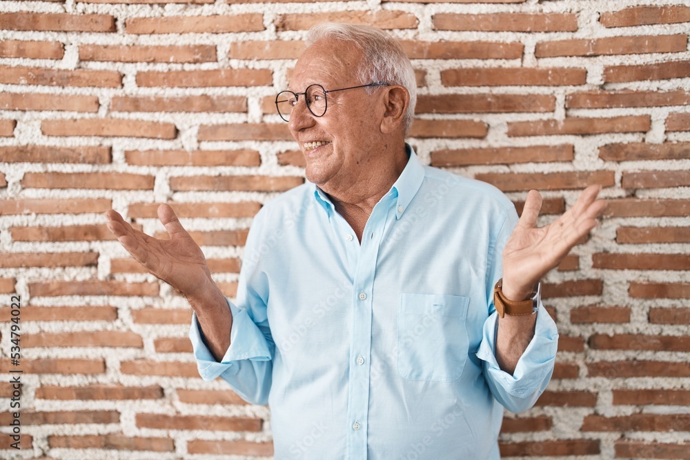 Senior man with grey hair standing over bricks wall smiling showing both hands open palms, presenting and advertising comparison and balance Senior man with grey hair standing over bricks wall smiling showing both hands open palms, presenting and advertising comparison and balance