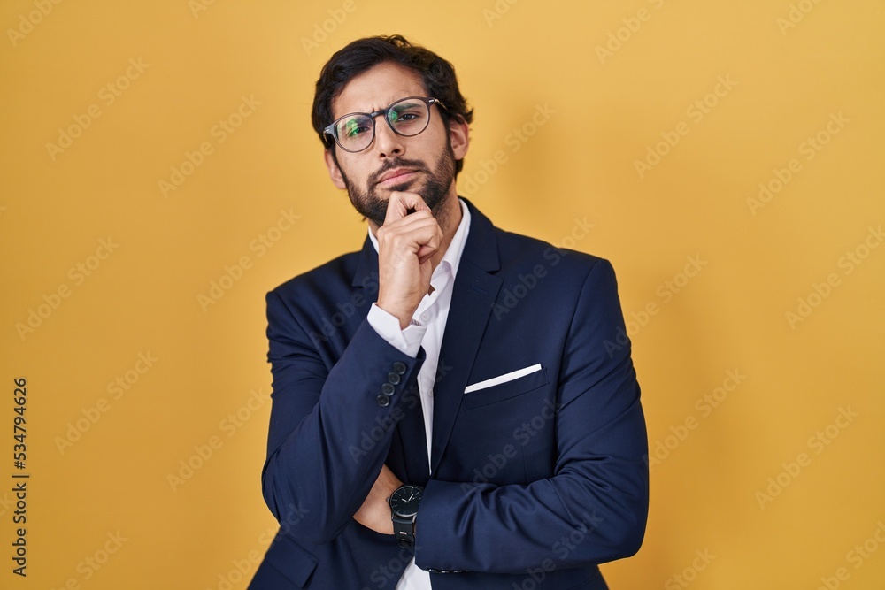 Handsome latin man standing over yellow background looking confident at the camera smiling with crossed arms and hand raised on chin. thinking positive. Handsome latin man standing over yellow background looking confident at the camera smiling with crossed arms and hand raised on chin. thinking positive.