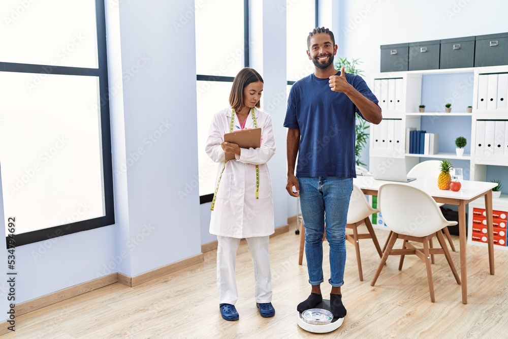 Fototapeta premium Dietitian at the clinic with client checking weight smiling happy and positive, thumb up doing excellent and approval sign