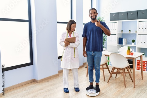 Dietitian at the clinic with client checking weight smiling happy and positive, thumb up doing excellent and approval sign © Krakenimages.com