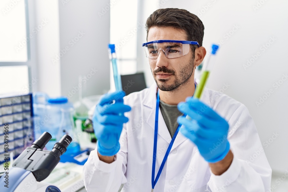 Young hispanic man wearing scientist uniform holding test tubes at laboratory Young hispanic man wearing scientist uniform holding test tubes at laboratory