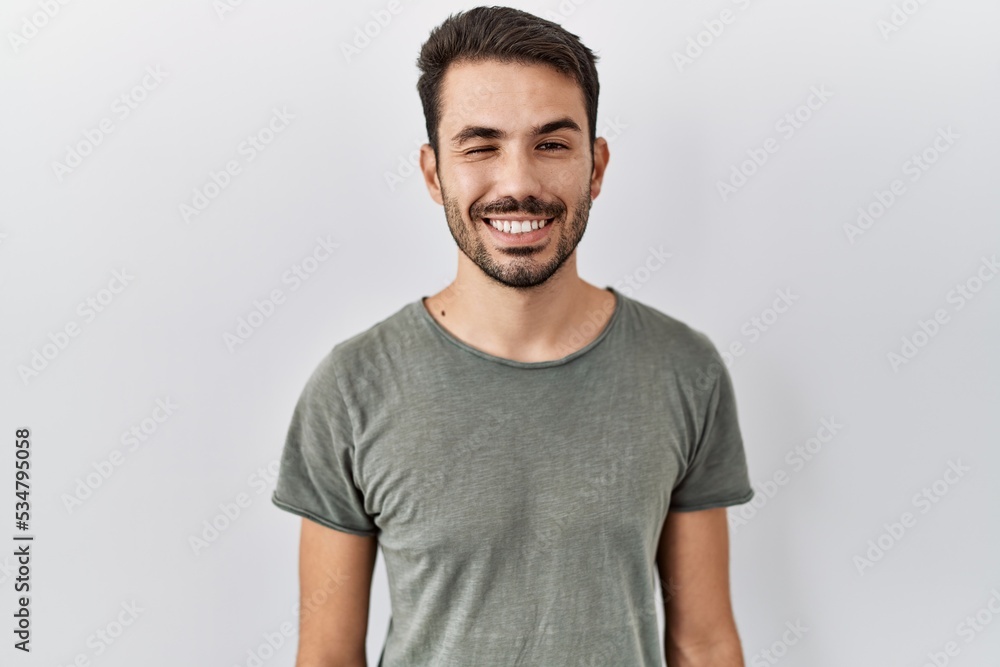 © Krakenimages.com - Young hispanic man with beard wearing casual t shirt over white background winking looking at the camera with sexy expression, cheerful and happy face.
