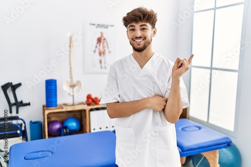 Young arab man working at pain recovery clinic with a big smile on face  pointing with hand and finger to the side looking at the camera.
