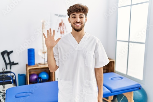 Young arab man working at pain recovery clinic showing and pointing up with fingers number five while smiling confident and happy.