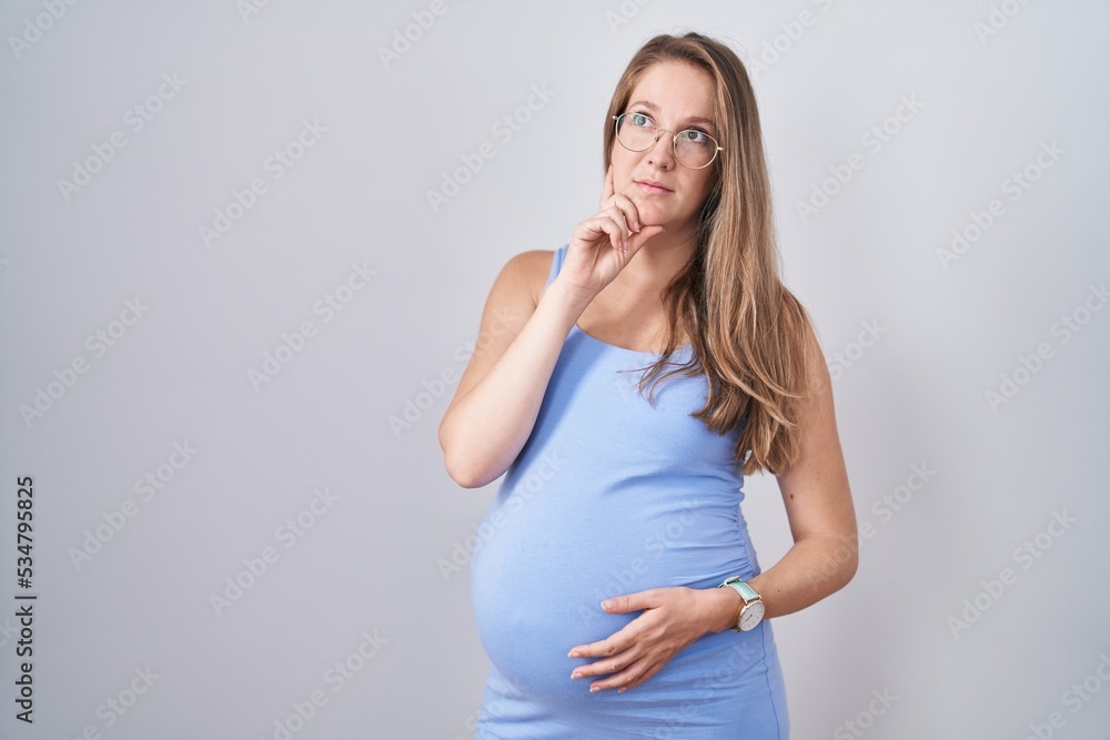 Young pregnant woman standing over white background with hand on chin thinking about question, pensive expression. smiling with thoughtful face. doubt concept. Young pregnant woman standing over white background with hand on chin thinking about question, pensive expression. smiling with thoughtful face. doubt concept.