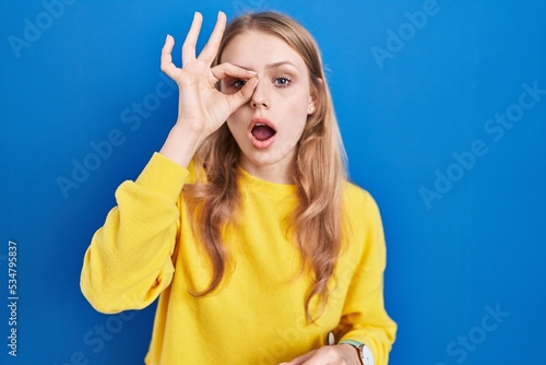 Young caucasian woman standing over blue background doing ok gesture shocked with surprised face, eye looking through fingers. unbelieving expression. © Krakenimages.com