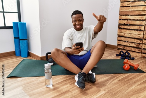 Young african man sitting on training mat at the gym using smartphone approving doing positive gesture with hand  thumbs up smiling and happy for success. winner gesture.
