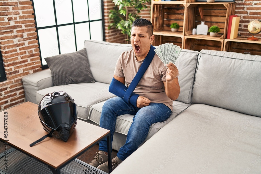 Hispanic young man wearing arm on sling holding usa dollars banknotes angry and mad screaming frustrated and furious, shouting with anger. rage and aggressive concept. Hispanic young man wearing arm on sling holding usa dollars banknotes angry and mad screaming frustrated and furious, shouting with anger. rage and aggressive concept.