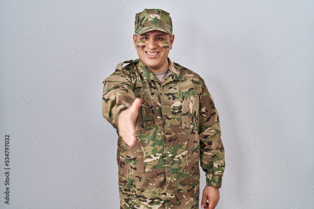 Hispanic young man wearing camouflage army uniform smiling friendly offering handshake as greeting and welcoming. successful business. Hispanic young man wearing camouflage army uniform smiling friendly offering handshake as greeting and welcoming. successful business.