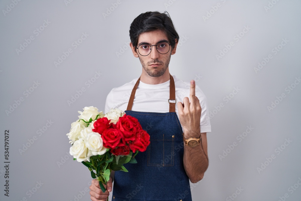 Young hispanic man holding bouquet of white and red roses showing middle finger, impolite and rude fuck off expression Young hispanic man holding bouquet of white and red roses showing middle finger, impolite and rude fuck off expression