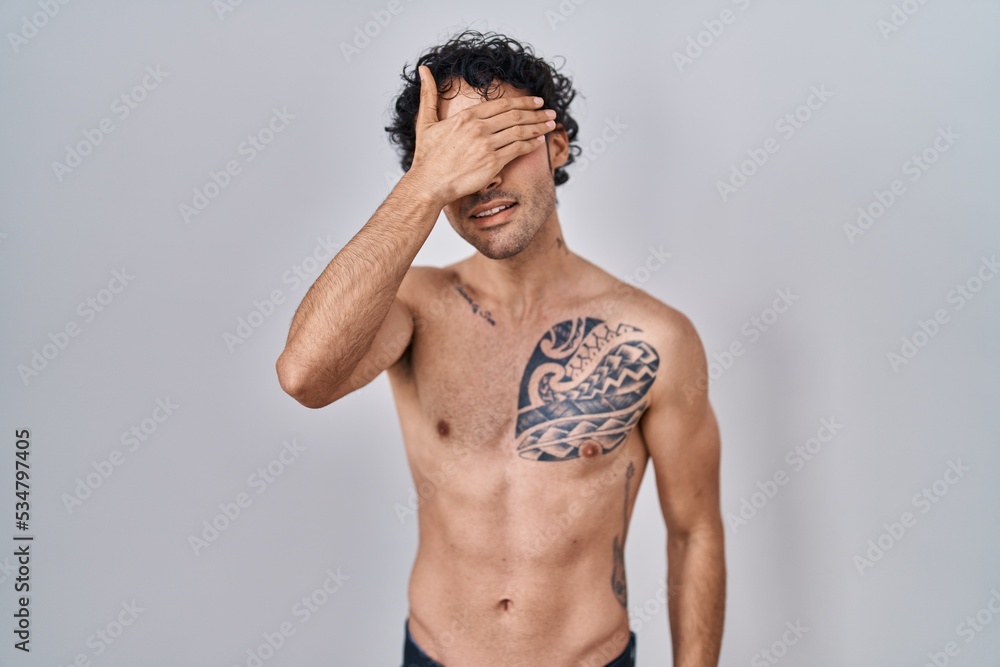 Hispanic man standing shirtless smiling and laughing with hand on face covering eyes for surprise. blind concept. Hispanic man standing shirtless smiling and laughing with hand on face covering eyes for surprise. blind concept.