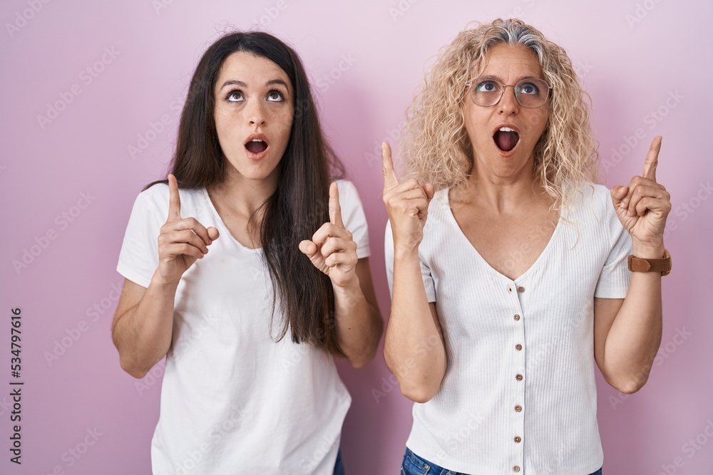 Mother and daughter standing together over pink background amazed and surprised looking up and pointing with fingers and raised arms. Mother and daughter standing together over pink background amazed and surprised looking up and pointing with fingers and raised arms.