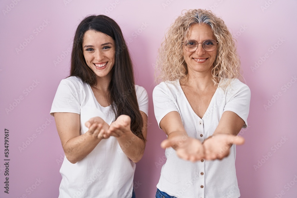 Mother and daughter standing together over pink background smiling with hands palms together receiving or giving gesture. hold and protection Mother and daughter standing together over pink background smiling with hands palms together receiving or giving gesture. hold and protection