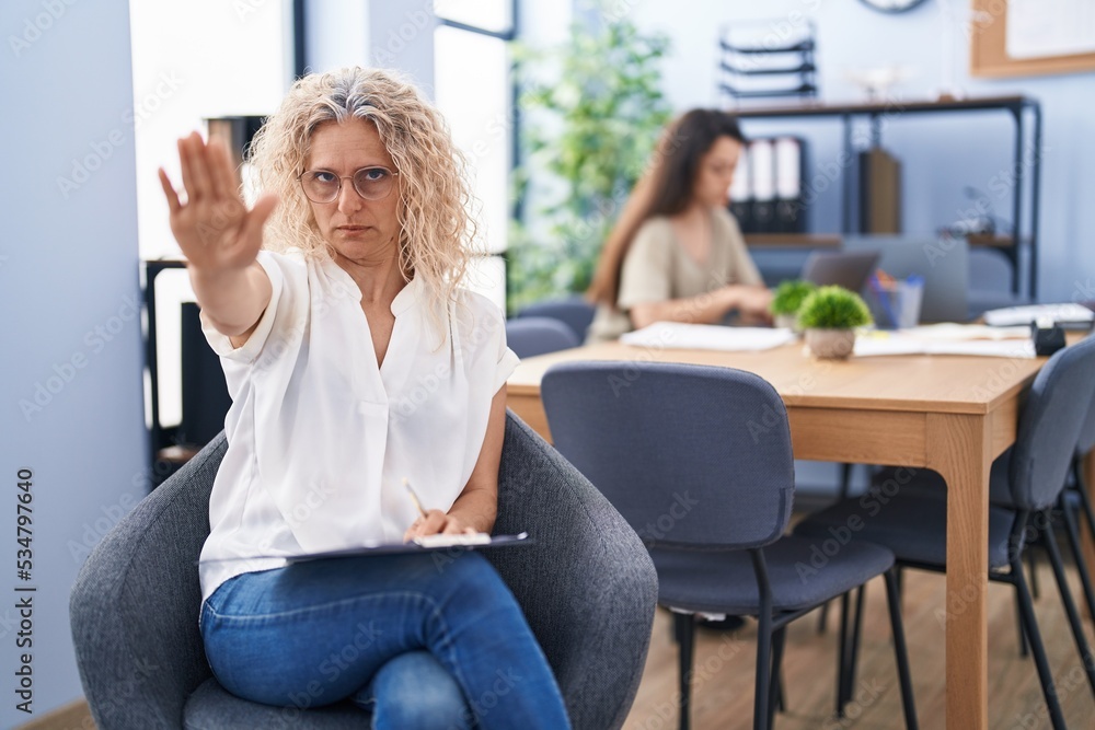 Middle age woman working at the office holding clipboard with open hand doing stop sign with serious and confident expression, defense gesture