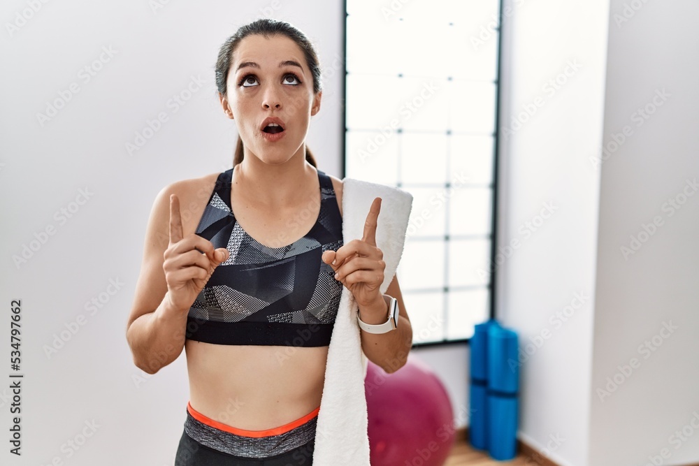 Young brunette woman wearing sportswear and towel at the gym amazed and surprised looking up and pointing with fingers and raised arms. Young brunette woman wearing sportswear and towel at the gym amazed and surprised looking up and pointing with fingers and raised arms.