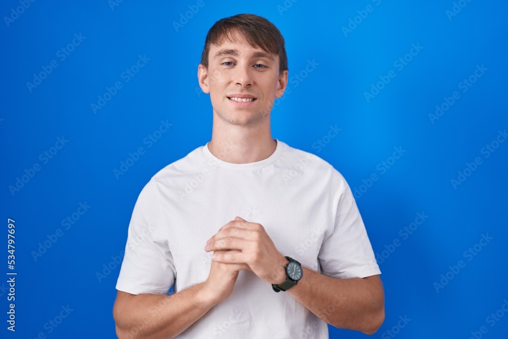 Caucasian blond man standing over blue background hands together and fingers crossed smiling relaxed and cheerful. success and optimistic Caucasian blond man standing over blue background hands together and fingers crossed smiling relaxed and cheerful. success and optimistic