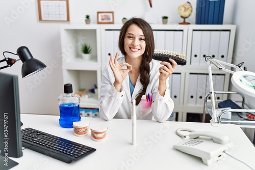 Young dentist woman comparing teeth whitening doing ok sign with fingers, smiling friendly gesturing excellent symbol
