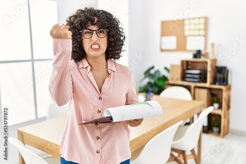 Young middle eastern woman wearing business style at office angry and mad raising fist frustrated and furious while shouting with anger. rage and aggressive concept.