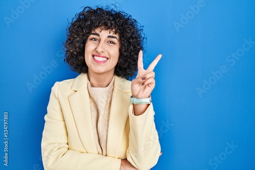 Young brunette woman with curly hair standing over blue background smiling with happy face winking at the camera doing victory sign with fingers. number two.