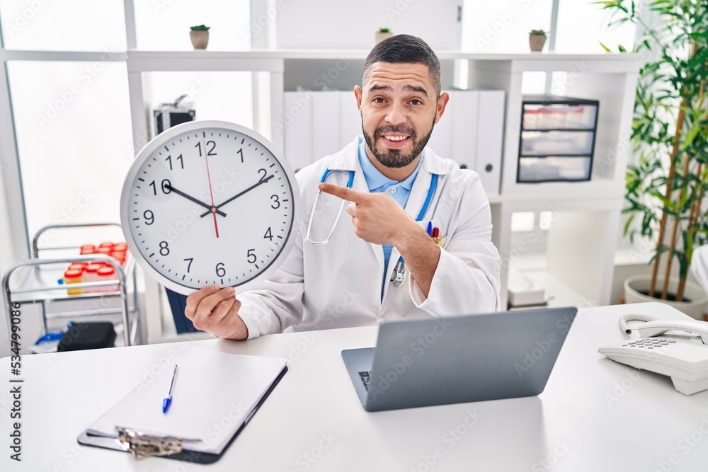 Hispanic doctor man holding clock at the clinic smiling happy pointing with hand and finger Hispanic doctor man holding clock at the clinic smiling happy pointing with hand and finger