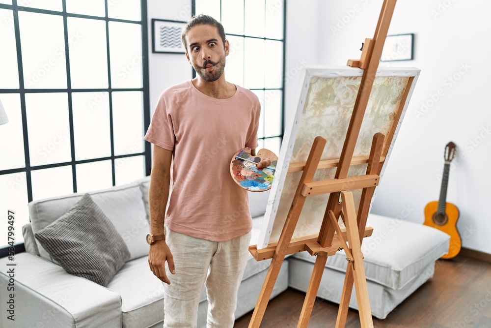 Young hispanic man with beard painting on canvas at home making fish face with lips, crazy and comical gesture. funny expression.