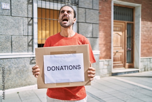 Young hispanic man holding donations box for charity outdoors angry and mad screaming frustrated and furious, shouting with anger looking up.