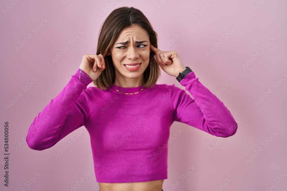 Hispanic woman standing over pink background covering ears with fingers with annoyed expression for the noise of loud music. deaf concept. Hispanic woman standing over pink background covering ears with fingers with annoyed expression for the noise of loud music. deaf concept.