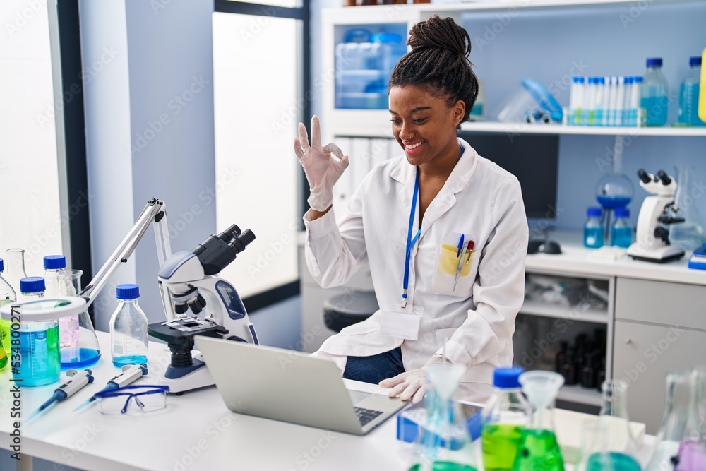 © Krakenimages.com - Young african american with braids working at scientist laboratory with laptop doing ok sign with fingers, smiling friendly gesturing excellent symbol