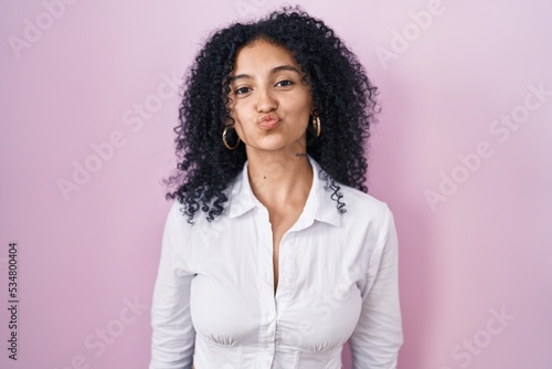 Hispanic woman with curly hair standing over pink background looking at the camera blowing a kiss on air being lovely and sexy. love expression. © Krakenimages.com