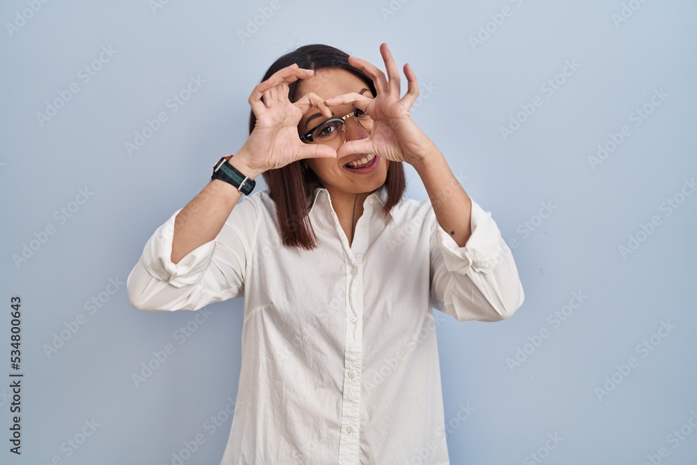Young hispanic woman standing over white background doing heart shape with hand and fingers smiling looking through sign Young hispanic woman standing over white background doing heart shape with hand and fingers smiling looking through sign