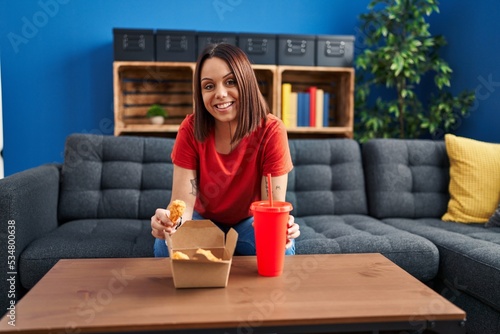 Young beautiful hispanic woman eating fried chicken sitting on sofa at home