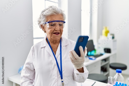 Senior grey-haired woman wearing scientist uniform using smartphone at laboratory