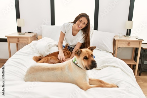 Young hispanic woman playing with dogs sitting on bed at bedroom