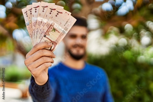 Young arab man smiling confident holding riyal banknotes at park photo