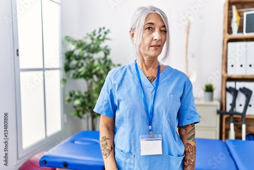 Middle age grey-haired woman wearing physiotherapist uniform at medical clinic winking looking at the camera with sexy expression, cheerful and happy face.