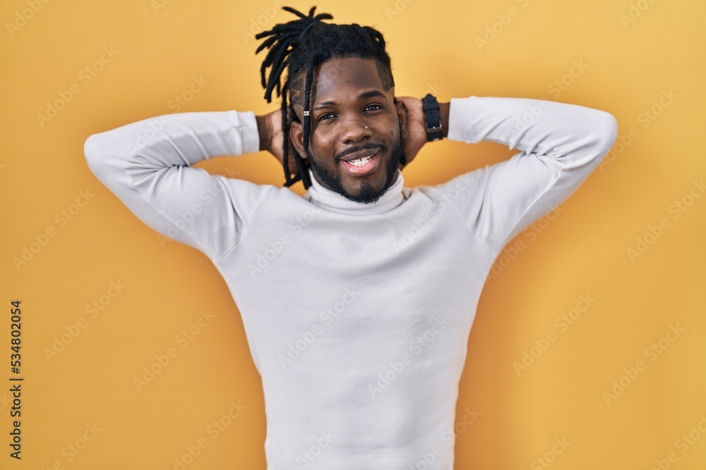 African man with dreadlocks wearing turtleneck sweater over yellow background relaxing and stretching, arms and hands behind head and neck smiling happy African man with dreadlocks wearing turtleneck sweater over yellow background relaxing and stretching, arms and hands behind head and neck smiling happy
