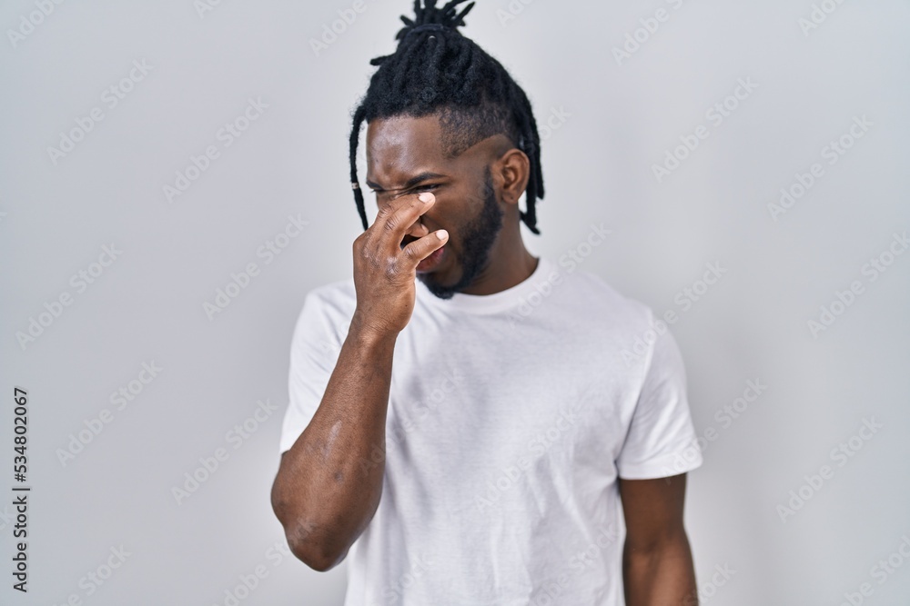 African man with dreadlocks wearing casual t shirt over white background smelling something stinky and disgusting, intolerable smell, holding breath with fingers on nose. bad smell African man with dreadlocks wearing casual t shirt over white background smelling something stinky and disgusting, intolerable smell, holding breath with fingers on nose. bad smell