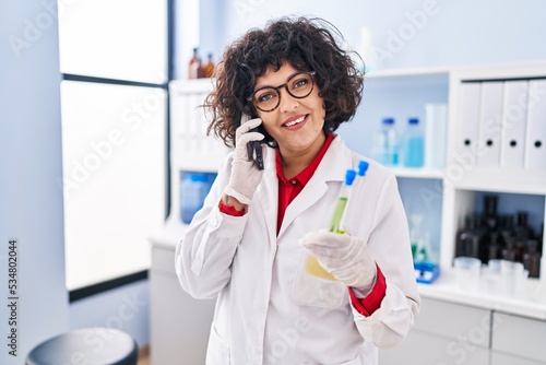 Young beautiful hispanic woman scientist talking on smartphone holding test tube at laboratory