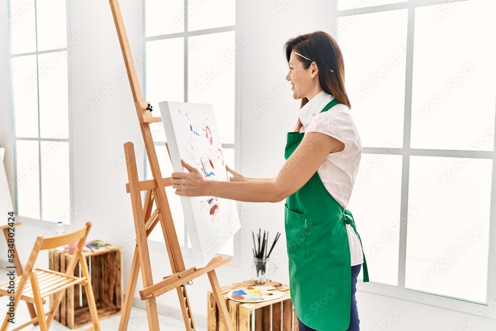 Middle age hispanic woman smiling confident holding canvas draw at art studio