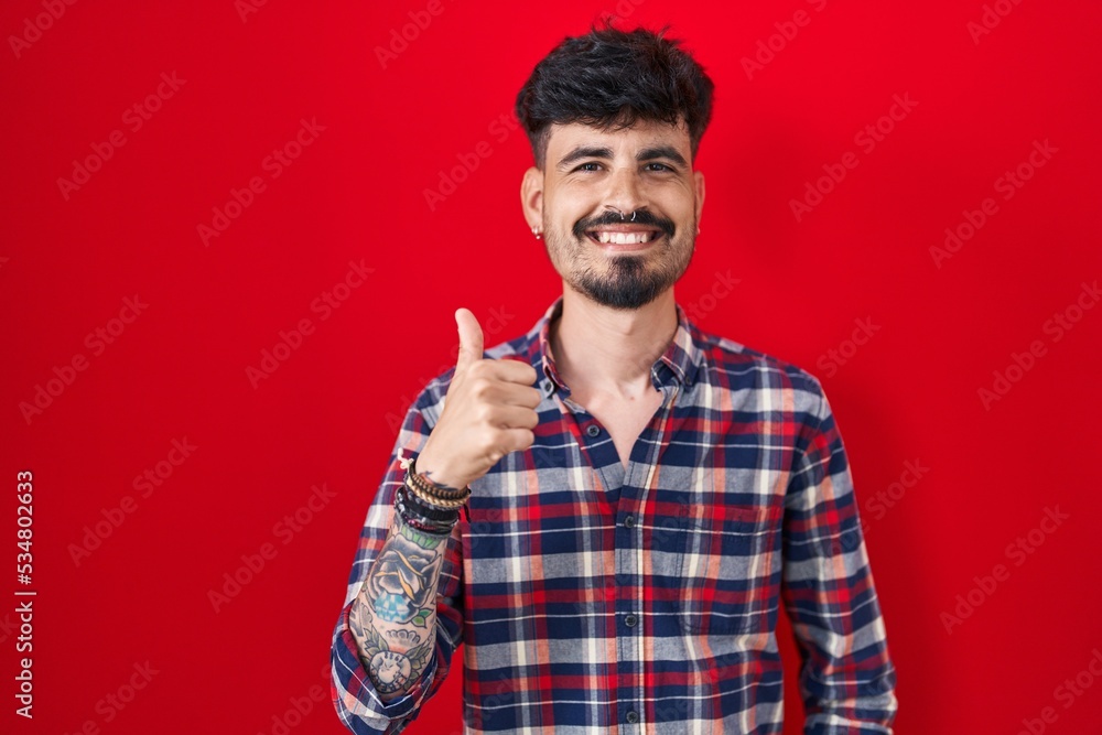 Young hispanic man with beard standing over red background doing happy thumbs up gesture with hand. approving expression looking at the camera showing success. Young hispanic man with beard standing over red background doing happy thumbs up gesture with hand. approving expression looking at the camera showing success.