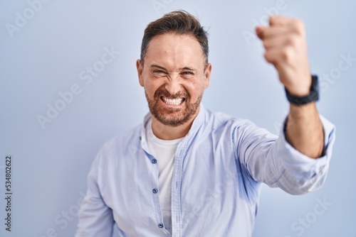 Middle age caucasian man standing over blue background angry and mad raising fist frustrated and furious while shouting with anger. rage and aggressive concept.