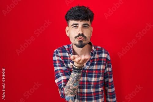 Young hispanic man with beard standing over red background looking at the camera blowing a kiss with hand on air being lovely and sexy. love expression.