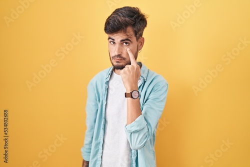 Young hispanic man with tattoos standing over yellow background pointing to the eye watching you gesture, suspicious expression