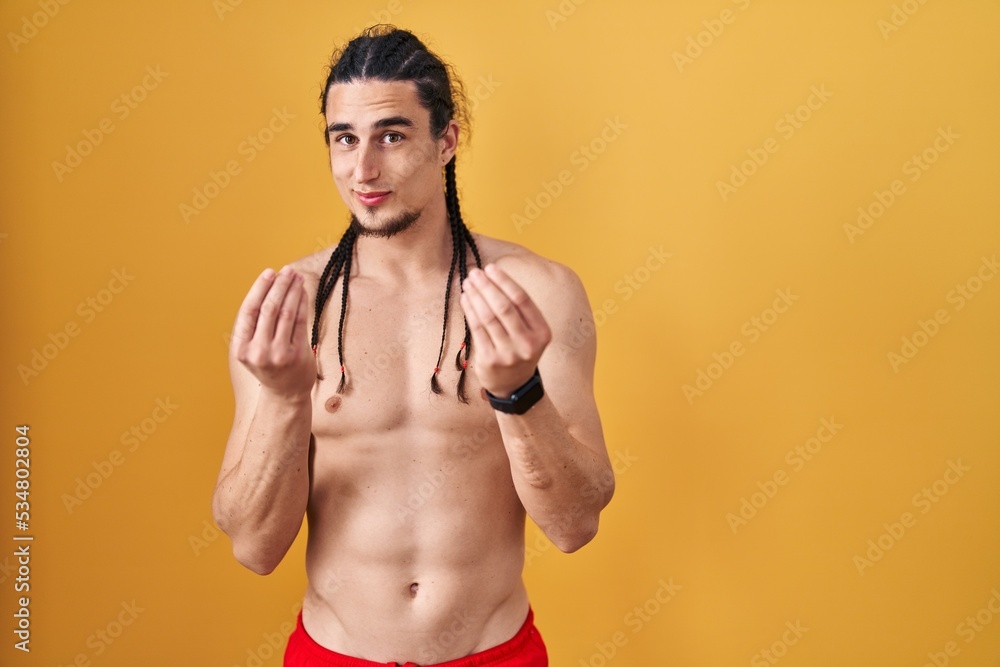 Hispanic man with long hair standing shirtless over yellow background doing money gesture with hands, asking for salary payment, millionaire business