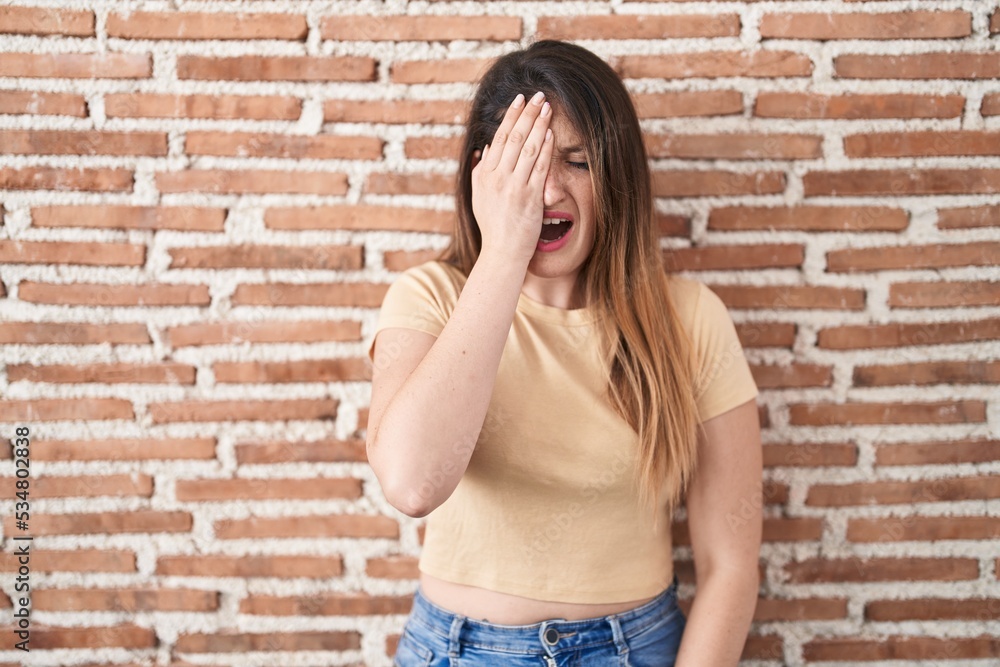 Young brunette woman standing over bricks wall yawning tired covering half face, eye and mouth with hand. face hurts in pain. Young brunette woman standing over bricks wall yawning tired covering half face, eye and mouth with hand. face hurts in pain.