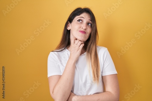 Young brunette woman standing over yellow background with hand on chin thinking about question, pensive expression. smiling with thoughtful face. doubt concept.