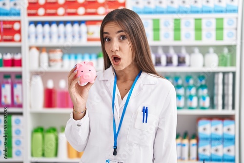 Young brunette woman working at pharmacy drugstore holding piggy bank scared and amazed with open mouth for surprise, disbelief face